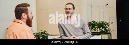 imprenditore ispirato in abiti casual e occhiali da vista ascoltando un partner commerciale barbuto che parla di progetto di avvio nella lobby di un ufficio moderno Foto Stock