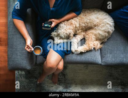 Vista dall'alto della donna sul divano che tiene il caffè e del telefono con il cane in braccio. Foto Stock