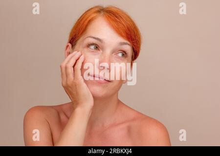 Ritratto di una donna di mezza età caucasica corta con lentiggini e capelli rossastri che tengono le mani sul mento su sfondo grigio guardando in alto Foto Stock