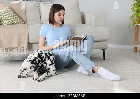 Bella giovane donna che legge un libro e il suo adorabile cane dalmata sul pavimento a casa. Adorabile animale domestico Foto Stock