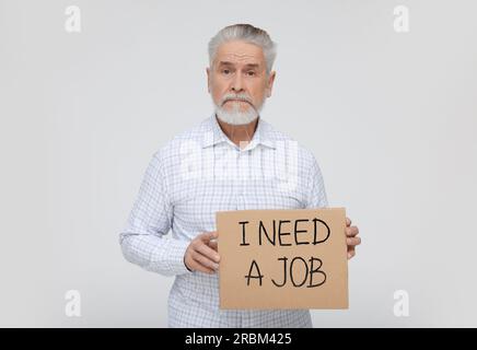 Un uomo anziano disoccupato con un cartello di cartone con la frase i Need A Job su sfondo bianco Foto Stock