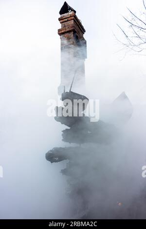 Resti di una casa dopo un incendio. Il camino in mattoni della stufa e le tavole carbonizzate sono avvolti da un fumo denso. Sfondo verticale con vista di Foto Stock