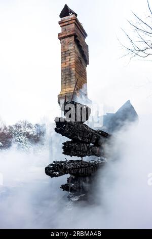 Resti di una casa dopo un incendio. Il camino in mattoni della stufa e le tavole carbonizzate sono avvolti da un fumo denso. Sfondo verticale con vista di Foto Stock