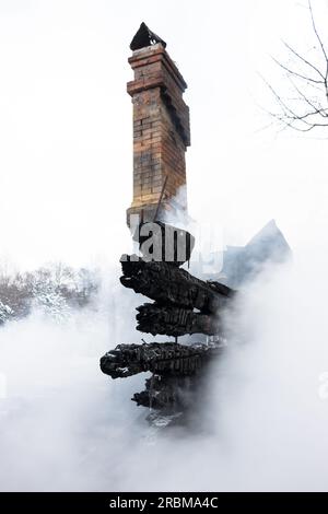 Resti di una casa dopo un incendio. Il camino in mattoni della stufa e le tavole carbonizzate sono avvolti da un fumo denso. Sfondo verticale con vista di Foto Stock