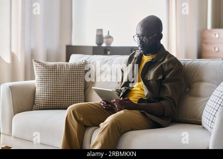 Giovane freelance afro-americano con un tablet seduto su un comodo e morbido divano in salotto e guardando i dati online Foto Stock