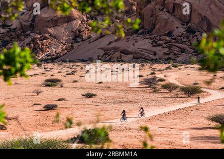 Diversi ciclisti su un sentiero sabbioso tra le rocce granitiche del gruppo Spitzkoppe Inselberg in Namibia, Africa Foto Stock