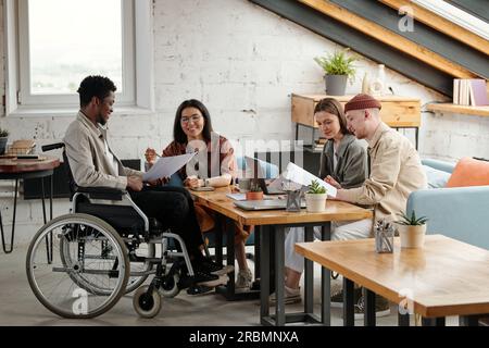 Due coppie di dipendenti che lavorano in piccoli gruppi per posto di lavoro, mentre economista donna che indica un documento tenuto da un collega maschio in sedia a rotelle Foto Stock