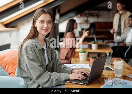 Giovane analista o economista sorridente che guarda la fotocamera mentre si trova sul posto di lavoro davanti al laptop e lavora con i dati finanziari online Foto Stock