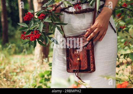 Primo piano della borsa in pelle marrone fatta a mano. Donna elegante che indossa abiti bianchi, cintura, borsa rustica con bacche rosse all'aperto. Donna boho informale Foto Stock