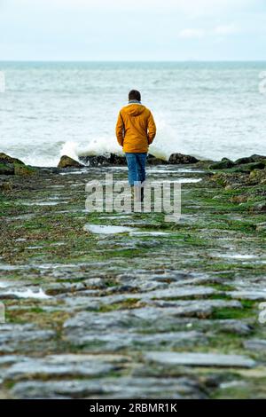 Donna con giacca arancione e pantaloni blu che cammina verso il mare. Foto Stock