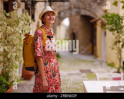 Giovane donna asiatica in vestito rosso e zaino cammina e guarda le strette stradine della città vecchia. Turismo, vacanza e scoperta concetto, viaggiatore femminile in visita sud Europa, isola di Rodi Grecia Foto Stock