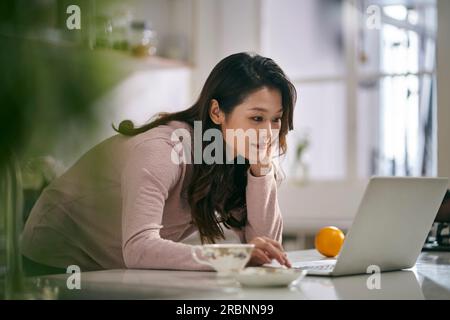 giovane donna d'affari asiatica che lavora in cucina a casa utilizzando un computer portatile Foto Stock