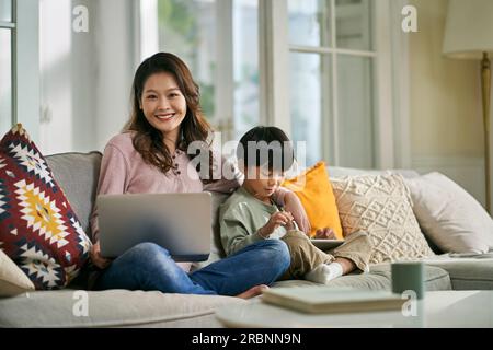 bella madre asiatica e figlio di cinque anni seduto sul divano di famiglia a casa guardando la macchina fotografica sorridente Foto Stock