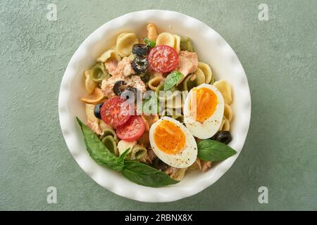 Insalata di pasta italiana. Orecchiette con tonno, ciliegia di pomodoro, oliva, basilico e parmigiano in piatto su fondo di pietra verde o cemento. Trad Foto Stock