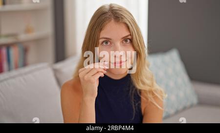 La giovane donna bionda seduta sul divano fa un gesto di chiusura della bocca a casa Foto Stock