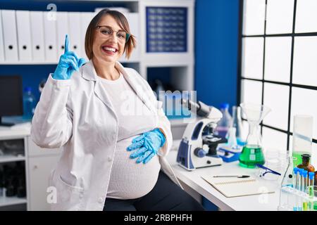 Donna incinta che lavora in un laboratorio di scienziati sorridendo stupita e sorpresa e puntando verso l'alto con le dita e le braccia sollevate. Foto Stock