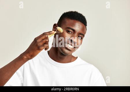 Ragazzo nero che usa il rullo facciale antigonfiamento giada per mantenere la pelle morbida e liscia. Bel giovane uomo afro-americano che mantiene la sua bellezza naturale con Foto Stock