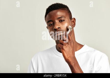 Bel ragazzo nero che usa il rullo per il viso che si sgonfia per mantenere la pelle morbida e liscia. Giovane afro-americano che mantiene la sua bellezza naturale con uno sci Foto Stock
