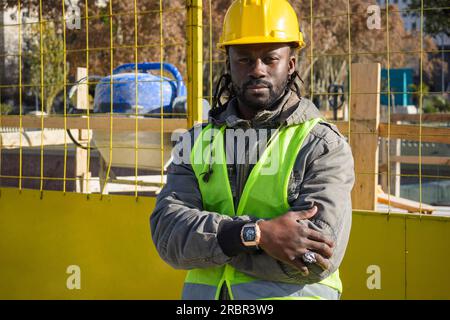 Ritratto di un giovane ingegnere africano che indossa un casco giallo, una giacca e un giubbotto, felice in piedi con le braccia incrociate che guardano la macchina fotografica, un armadietto di ingegneria Foto Stock
