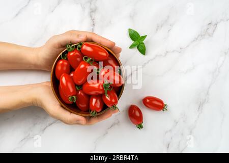 Ciotola di pomodori ciliegini Ornela freschi in una donna che passa su fondo di marmo. Le mani femminili tengono una ciotola di pomodori piccoli maturi rossi. Verdure biologiche. Foto Stock