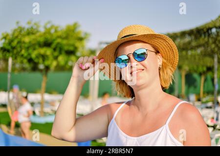 Felice giovane donna sorridente con cappello di paglia e occhiali da sole che si gode le vacanze estive. Ritratto di una bella donna europea che si gode la soleggiata giornata estiva. Foto Stock