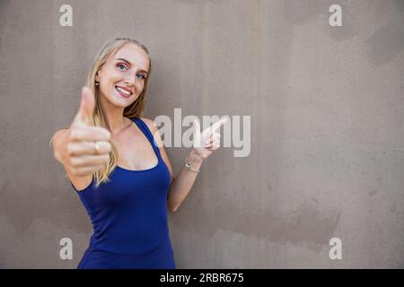 Una ragazza sorridente mostra il pollice verso l'alto e punta verso lo spazio sulla destra, pubblicando una foto con spazio per la copia, sfondo grigio Foto Stock