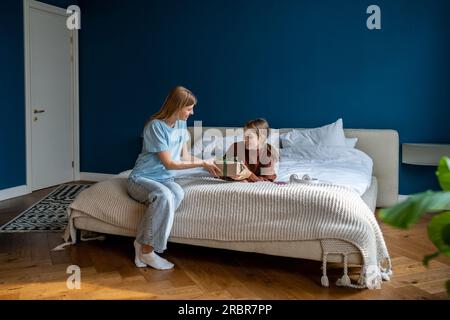 La madre premurosa si congratula con la figlia adolescente che si trova a letto e regala una confezione regalo per il compleanno Foto Stock