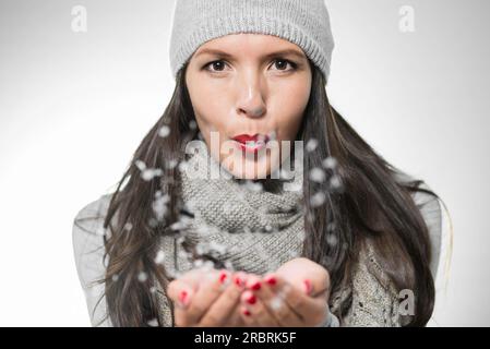 Attraente giovane donna bruna in caldo stile invernale che soffia fiocchi di neve o polvere magica dal palmo con labbra rosse e un divertito luccichio in lei Foto Stock