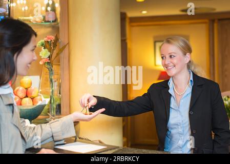 La receptionist dell'hotel sorridente e cordiale si trova dietro il banco del servizio nella hall di un hotel, prenotando con una cliente donna che le consegna le chiavi della camera Foto Stock