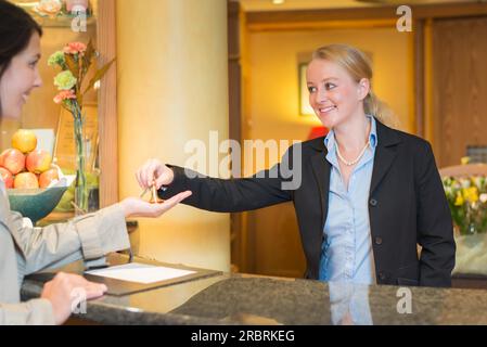 La receptionist dell'hotel sorridente e cordiale si trova dietro il banco del servizio nella hall di un hotel, prenotando con una cliente donna che le consegna le chiavi della camera Foto Stock