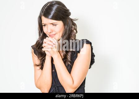 Sconvolta teary giovane donna con le mani giunte in angoscia in piedi con abbattuto gli occhi contro un bianco di sfondo per studio Foto Stock