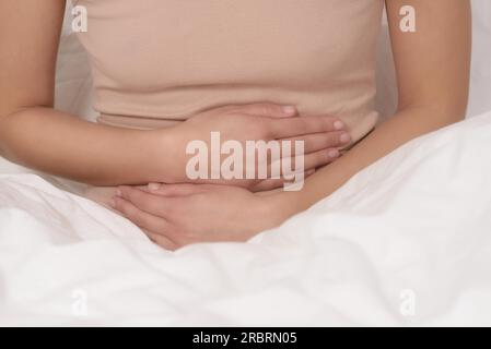 Donna con il suo mensile dolori mestruali stringendo il suo stomaco con le sue mani come ella diventa sottolineato dalle continue crampi mentre è seduto nel suo letto Foto Stock