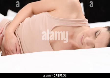 Donna con il suo mensile dolori mestruali stringendo il suo stomaco con le sue mani come ella diventa sottolineato dalle continue crampi mentre è seduto nel suo letto Foto Stock