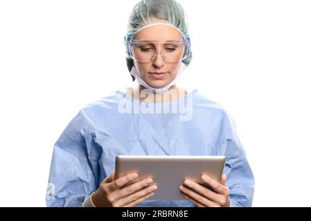 Attraente medico o chirurgo in scrub e camice chirurgico, occhiali e cappuccio che immettono le informazioni del paziente su un tablet computer, isolato su Foto Stock