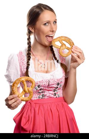 Felice e attraente giovane tedesca o bavarese in un dirndl che mangia pretzel tenendo due n le mani con la bocca spalancata per prendere un morso, isolata Foto Stock