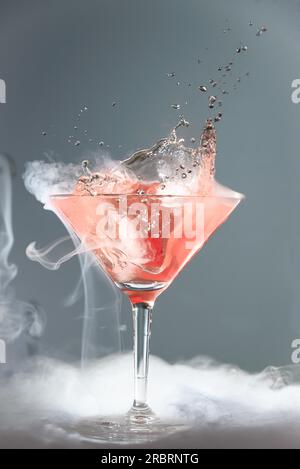 Cocktail martini fumanti in un bicchiere conico con vapori e gocce di ciliegia cadenti per un effetto drammatico su un grigio Foto Stock