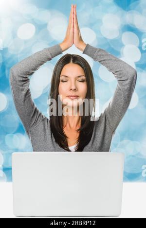 Capelli piuttosto lunghi giovane donna che medita con le mani sopra la testa e gli occhi chiusi davanti a un computer portatile, catturata in Studio su Abstract Blue Foto Stock