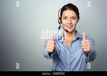 Attraente operatore di call center, assistente del servizio clienti o operatore di telemarketing che indossa una cuffia che guarda la fotocamera e tiene il pollice in alto Foto Stock