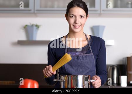 Giovane casalinga che prepara una sana pasta italiana in piedi alla stufa con un piatto pieno di spaghetti con verdure fresche in primo piano sorridente Foto Stock