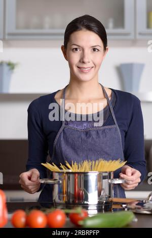 Giovane casalinga che prepara una sana pasta italiana in piedi alla stufa con un piatto pieno di spaghetti con verdure fresche in primo piano sorridente Foto Stock