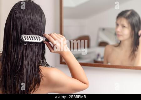 Giovane donna in piedi che guarda il suo riflesso nello specchio del bagno mentre si spazzola i capelli bagnati con una spazzola dopo averla lavata, vista da dietro Foto Stock