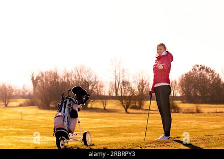 Giovane golfista con giacca e jeans che tiene un club mentre regala un pollice con il golf cart sul lato Foto Stock