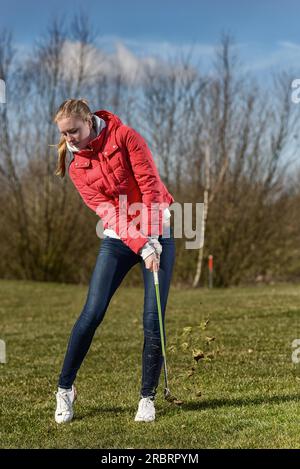 A piena lunghezza shot di un giovane giocatore di golf femminile, indossa una giacca rossa e Skinny Jeans, nella ruvida, appena dopo aver disputato una sfera Foto Stock