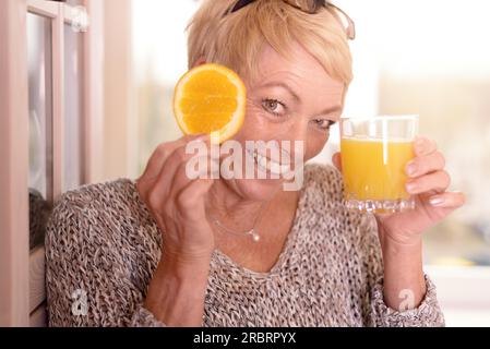 Sorridente e vivace donna bionda di mezza età che tiene una fetta d'arancia all'occhio mentre tiene un bicchiere di succo d'arancia appena spremuto nell'altro in A. Foto Stock