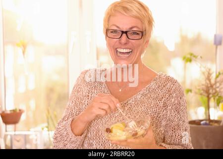 Vivace e attraente donna di mezza età che indossa gli occhiali che mangia una sana ciotola di Macedonia di frutta ricca di vitamine che guarda la fotocamera con un segnale acustico Foto Stock