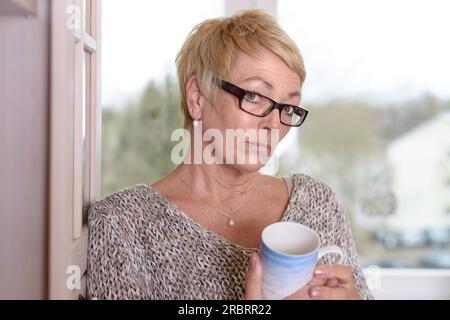 Primo piano donna seria del Medioevo che indossa occhiali da vista, con capelli biondi corti, che tiene una tazza mentre si appoggia alla parete e guarda la fotocamera Foto Stock