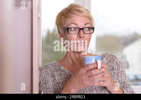 Primo piano donna seria del Medioevo che indossa occhiali da vista, con capelli biondi corti, che tiene una tazza mentre si appoggia alla parete e guarda la fotocamera Foto Stock