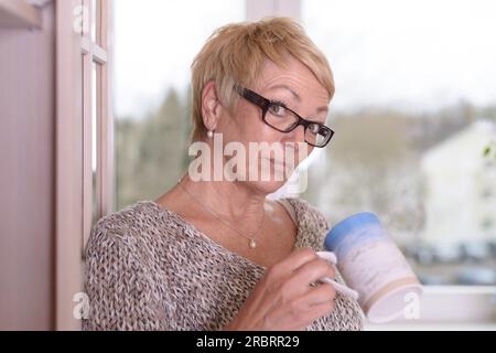 Primo piano donna seria del Medioevo che indossa occhiali da vista, con capelli biondi corti, che tiene una tazza mentre si appoggia alla parete e guarda la fotocamera Foto Stock