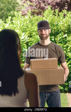 Donna rispondendo alla porta a un attraente barbuto deliveryman portante due scatole di cartone per la consegna, vista sulla sua spalla da dietro Foto Stock