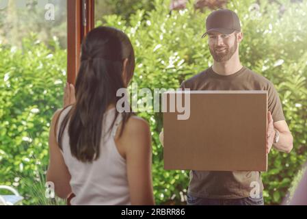 Donna rispondendo alla porta a un attraente barbuto deliveryman portante due scatole di cartone per la consegna, vista sulla sua spalla da dietro Foto Stock